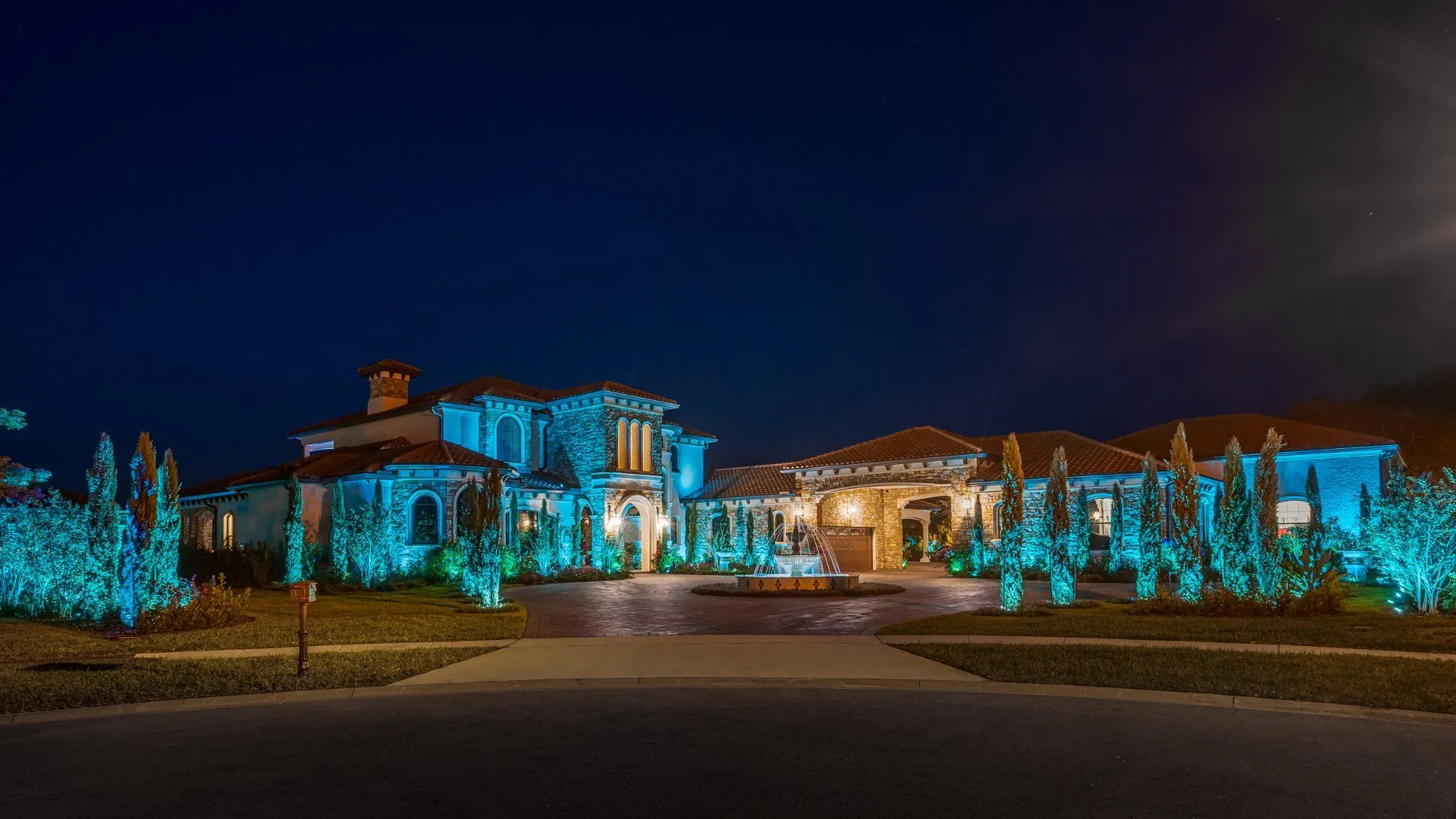 A large Mediterranean-style custom home photographed at dusk, its stone facade, tile roof and surrounding cypress trees lit by teal landscape uplighting, with a tiered fountain in the circular paver driveway.