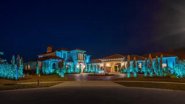 A large Mediterranean-style custom home photographed at dusk, its stone facade, tile roof and surrounding cypress trees lit by teal landscape uplighting, with a tiered fountain in the circular paver driveway.